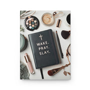 Black notebook with 'Wake. Pray. Slay.' text on a white surface with makeup items.